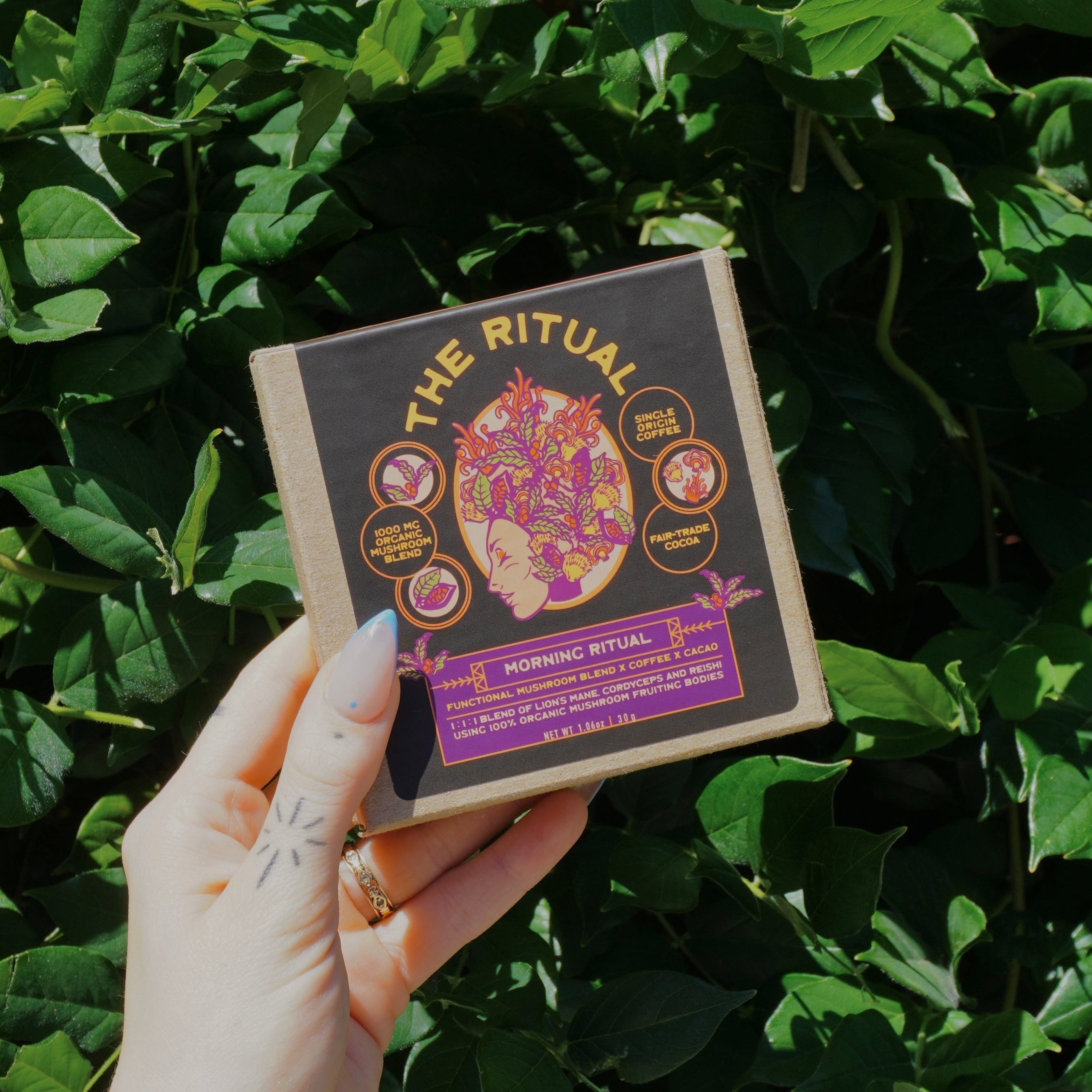 A hand holding a square product box labeled 'The Ritual' in front of a dense background of green ivy leaves.