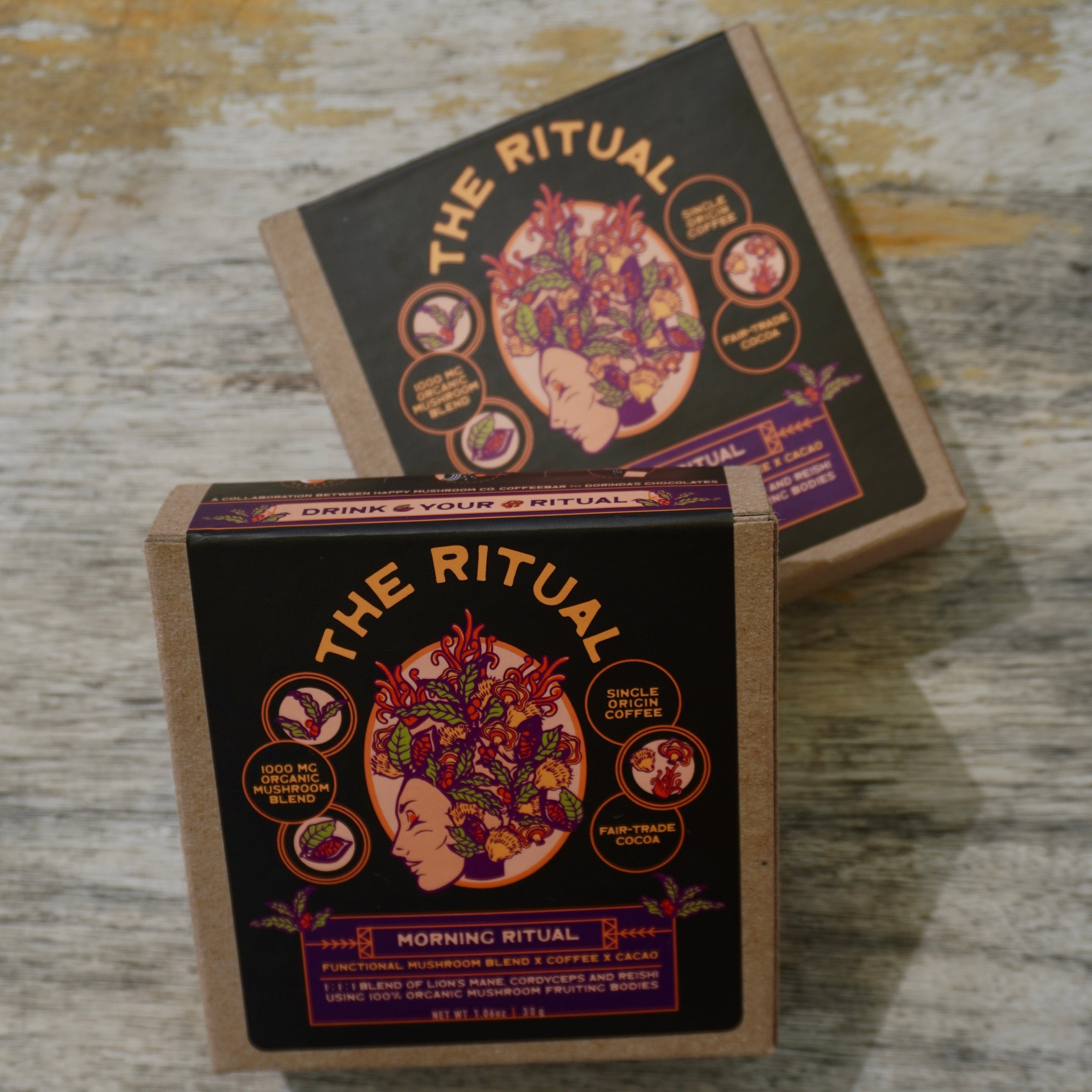 Two square boxes of 'The Ritual' Morning Ritual, featuring a dark black label with a colorful illustration of a woman's profile and botanical elements, resting on a rustic wooden surface.