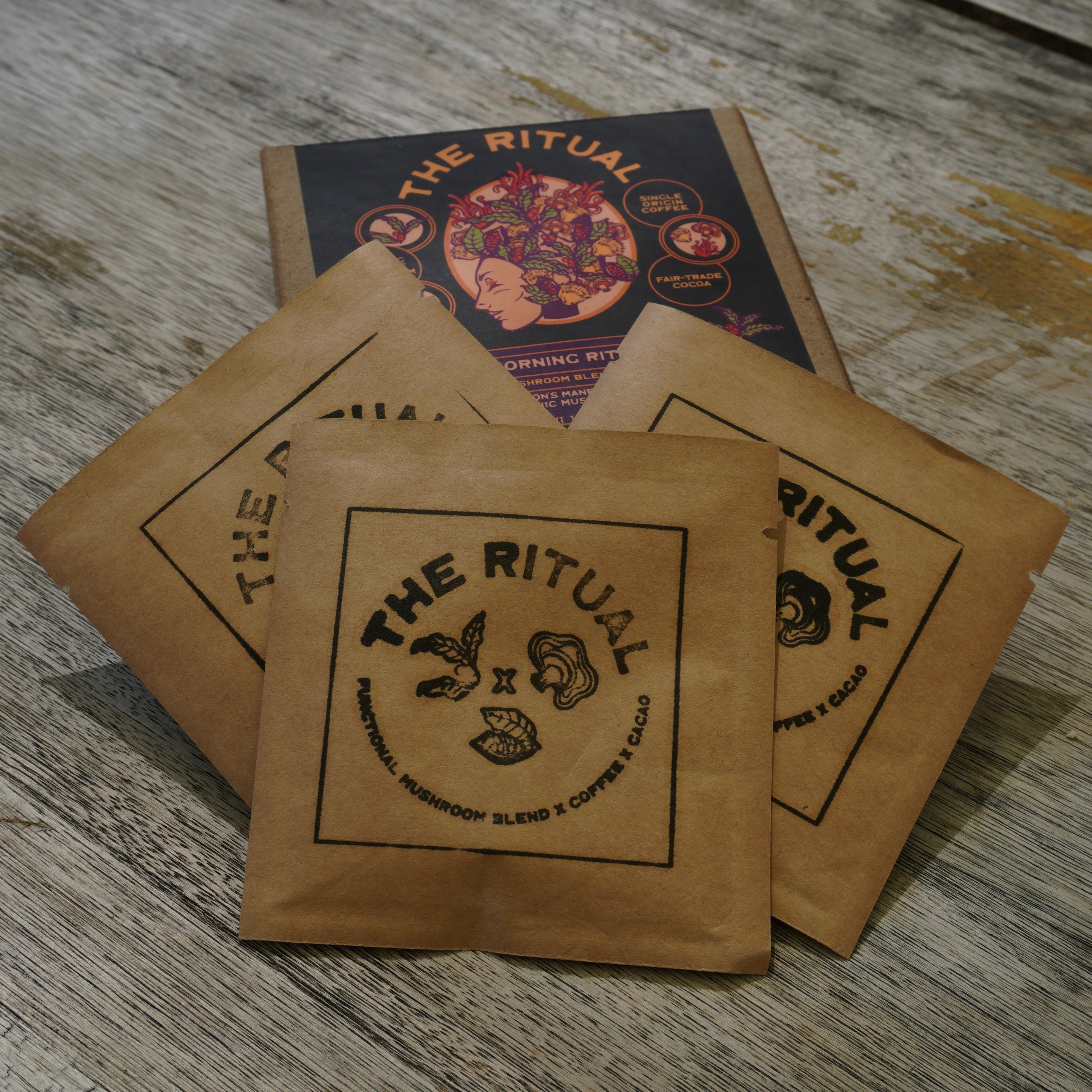 Three kraft paper coffee bags with black printed logos from 'The Ritual' are arranged on a rustic, weathered gray wooden tabletop, with a branded box visible in the background.