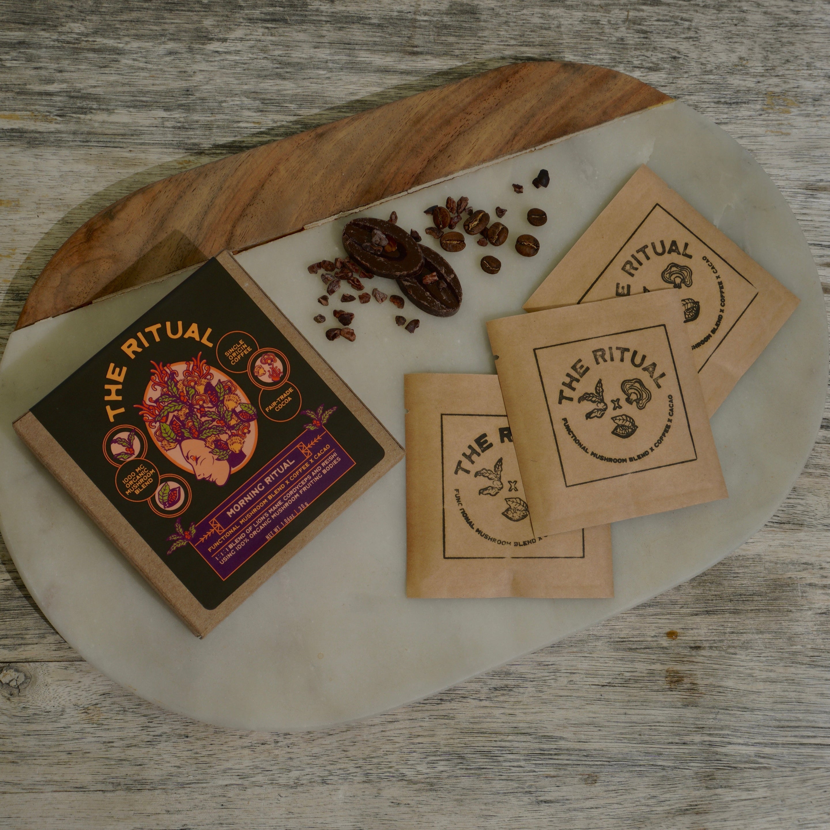 A styled flat-lay of 'The Ritual' brand product on a white oval tray with a wooden edge, set on a weathered gray wood surface. The product includes a dark brown illustrated box, three small kraft paper sachets, and scattered coffee beans and star anise.