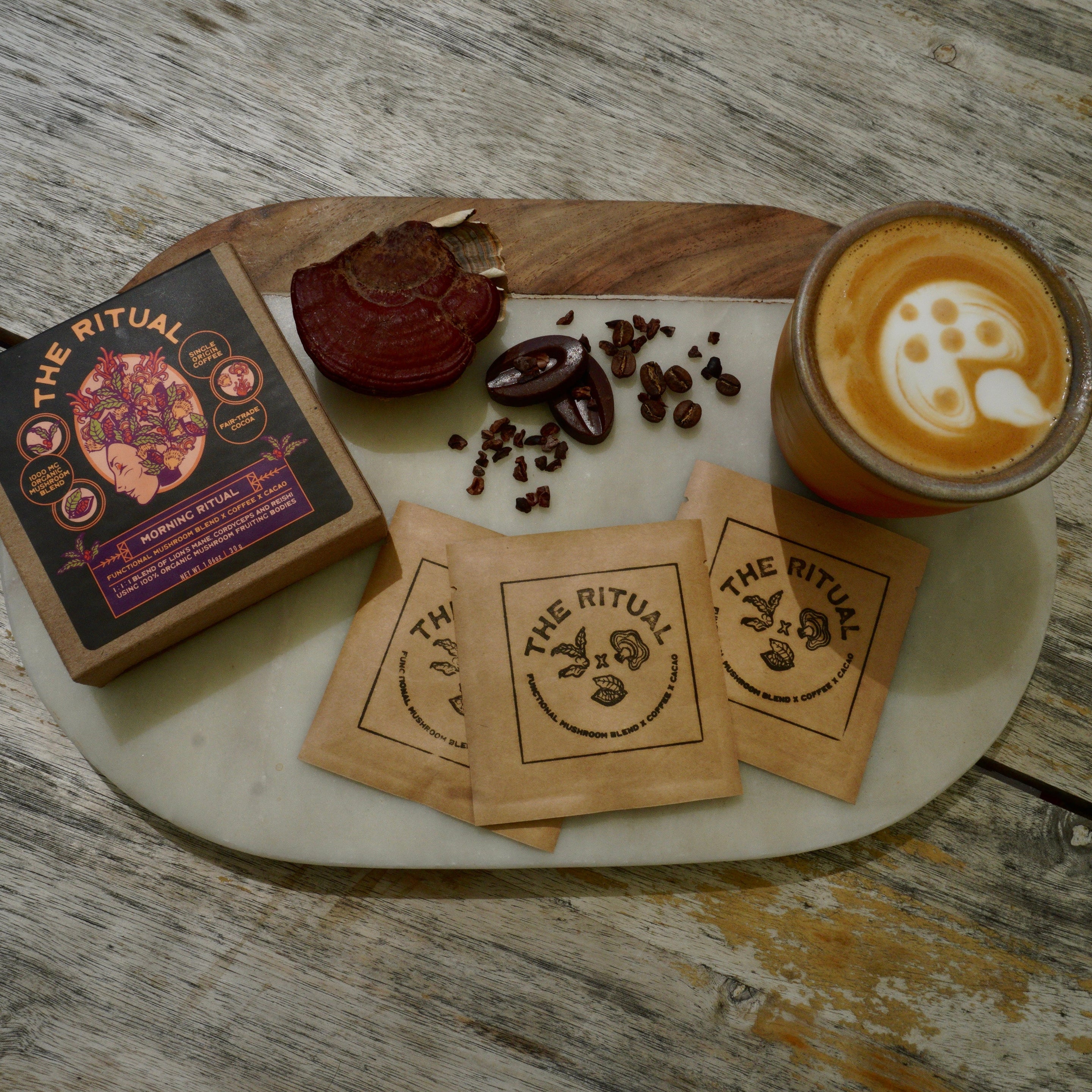 A styled flat-lay of 'The Ritual' mushroom coffee product on a rustic wooden table, featuring a coffee box, three branded paper sachets, loose coffee beans, a dried red reishi mushroom, and a glass of latte art coffee on a white tray.